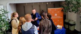 Zdjęcie przedstawia stoisko Generalnej Dyrekcji Dróg Krajowych i Autostrad (GDDKiA) na targach pracy organizowanych przez Politechnikę Śląską. Przedstawiciel instytucji prowadzi rozmowę z grupą zainteresowanych osób, które prawdopodobnie poszukują ofert pracy lub staży. Na stole rozmieszczone są materiały informacyjne, a w tle widoczny jest duży baner z logo GDDKiA oraz pełną nazwą organizacji. Dookoła stoiska znajdują się rośliny doniczkowe, co nadaje przestrzeni bardziej uporządkowany i przyjazny charakter.