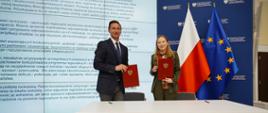 Dwie osoby stoją obok siebie i trzymają dokumenty. Pierwsza od prawej stoi minister Katarzyna Pełczyńska-Nałęcz.
