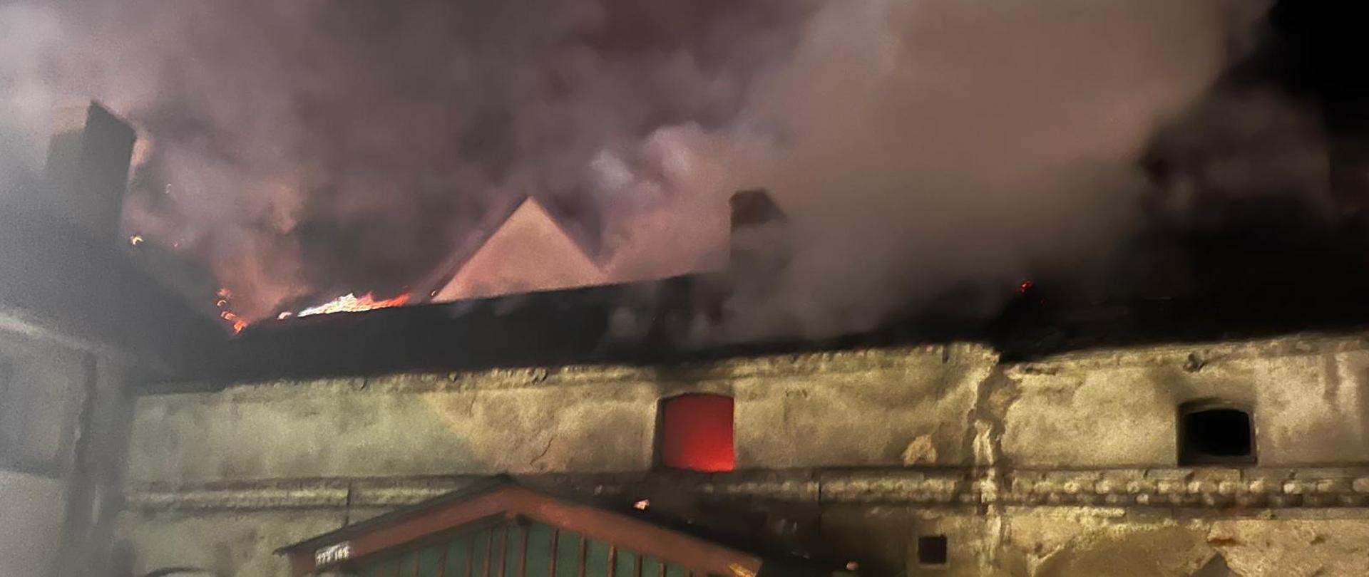 Fotografia przedstawia widok opuszczonego budynku w nocy. Na dachu widoczne płomienie oraz unoszący się dym.