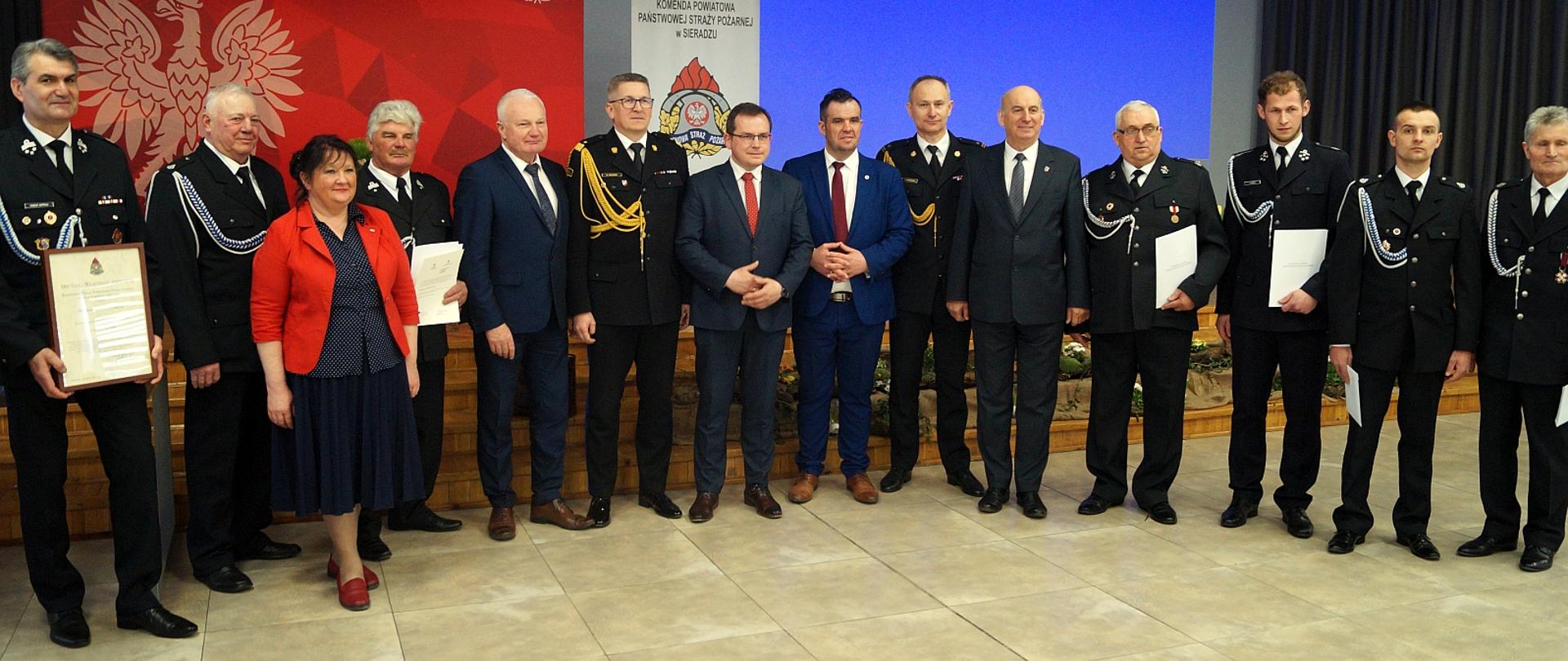 na zdjęciu strażacy oraz mężczyźni w garniturach i kobieta stojący w łuku na tle ściany