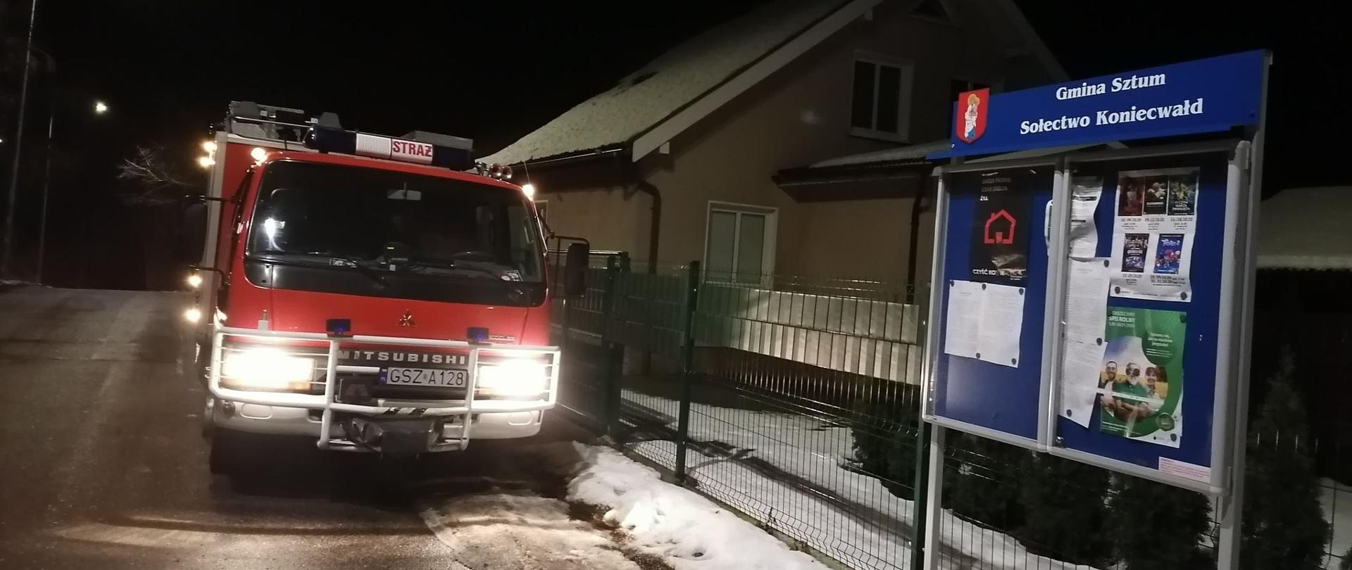 Na pierwszym planie stoi niebieska tablica informacyjna z herbem miasta, za nią samochód specjalny jednostki OSP Gościszewo. w tle stoi domek jednorodzinny. Pora nocna