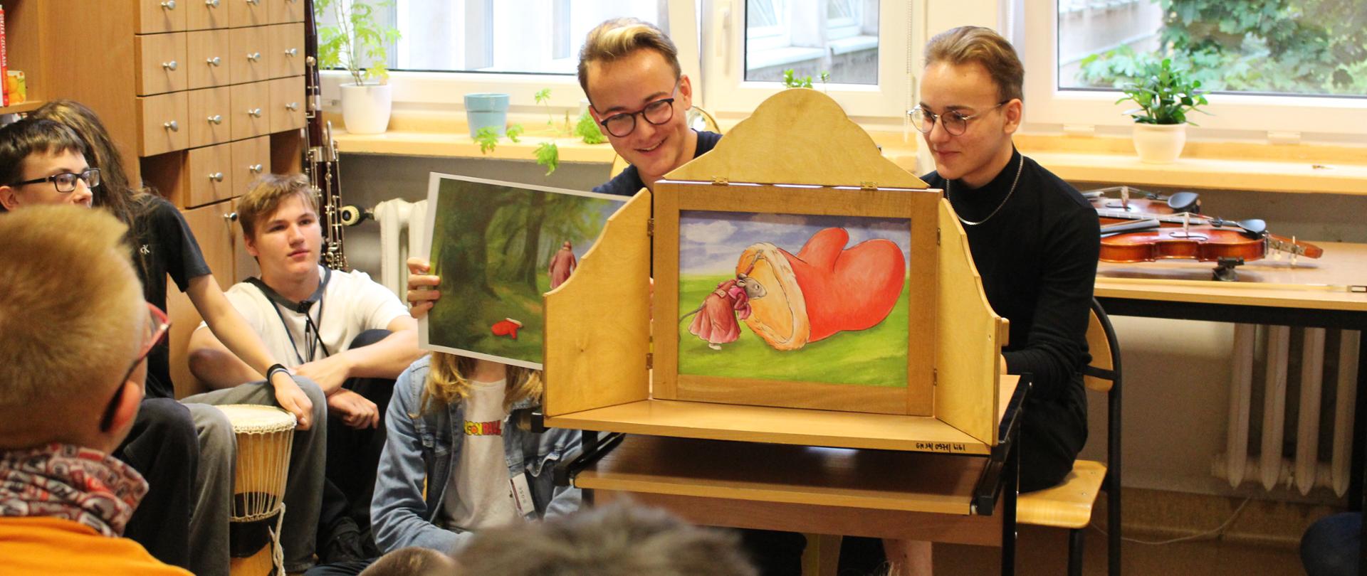 zdjęcie przedstawia uczniów prezentujących opowiadanie na rozkładanym ekranie na stoliku w sali edukacyjnej, inne ujęcie