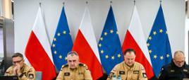 Trzech oficerów Państwowej Straży Pożarnej w żółtych mundurach służbowych i jeden w czarnym polarze siedzi przy stole konferencyjnym. Za nimi ustawiono flagi Polski i Unii Europejskiej. Na stole znajdują się mikrofony, napoje i dokumenty. Uczestnicy skupieni są na pracy przy laptopach.