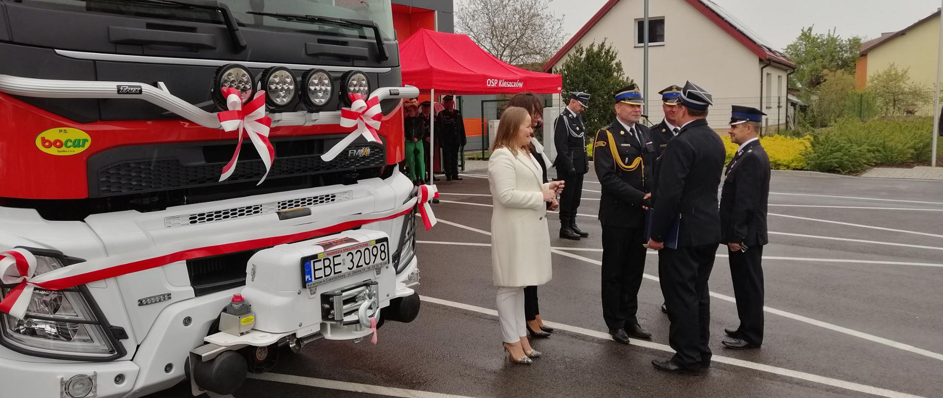 na głównym planie 2 kobiety i 4 strażaków, stojących obok samochodu pożarniczego, w tle budynki