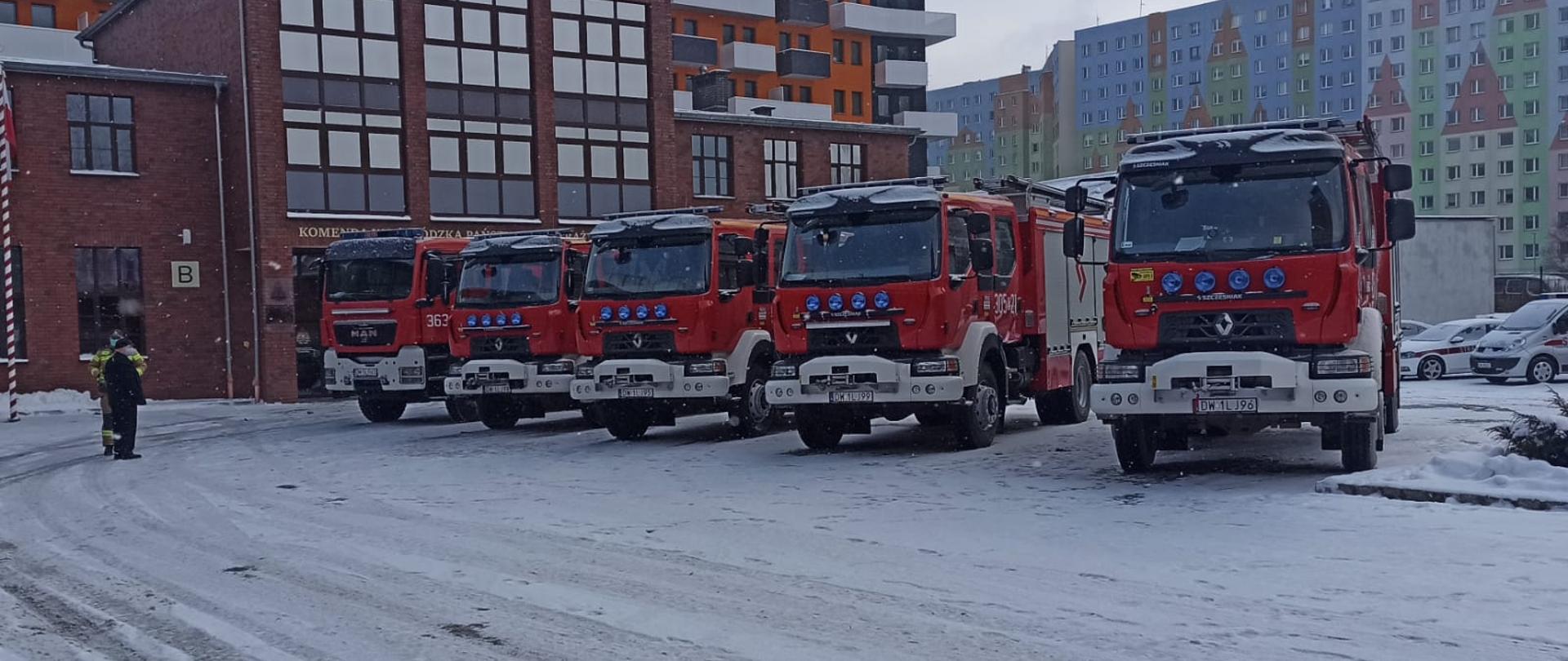 Po środku kadru stoi pięć czerwonych samochodów ciężarowych Państwowej Straży Pożarnej auta stoją na ośnieżonym placu w tle widać wielokolorowe budynki mieszkalne. Po lewej stronie widoczny masz na którym powiewa flaga Polski.