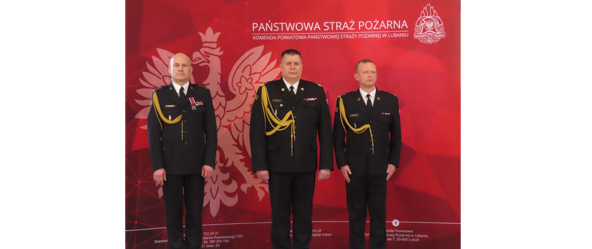 Komendant Wojewódzki, odchodzący Komendant Powiatowy i nowy Komendant Powiatowy stoją w garażu Komendy Powiatowej w Lubaniu na tle czerwonego baneru z logiem Państwowej Straży Pożarnej podczas uroczystości zdania i przyjęcia obowiązków Komendanta Powiatowego Państwowej Straży Pożarnej w Lubaniu. Komendanci są w mundurach wyjściowych.
