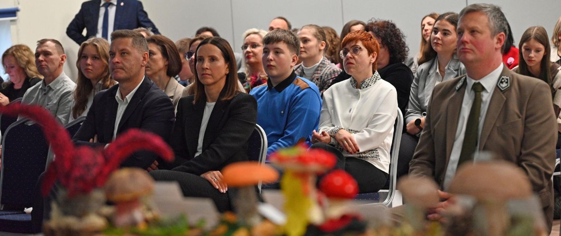 Uczestnicy uroczystości siedzący na widowni podczas finału konkursu. W pierwszym planie widoczne dekoracje w kształcie grzybów, w tle zgromadzeni goście – dorośli i młodzież – uważnie słuchający wystąpień.