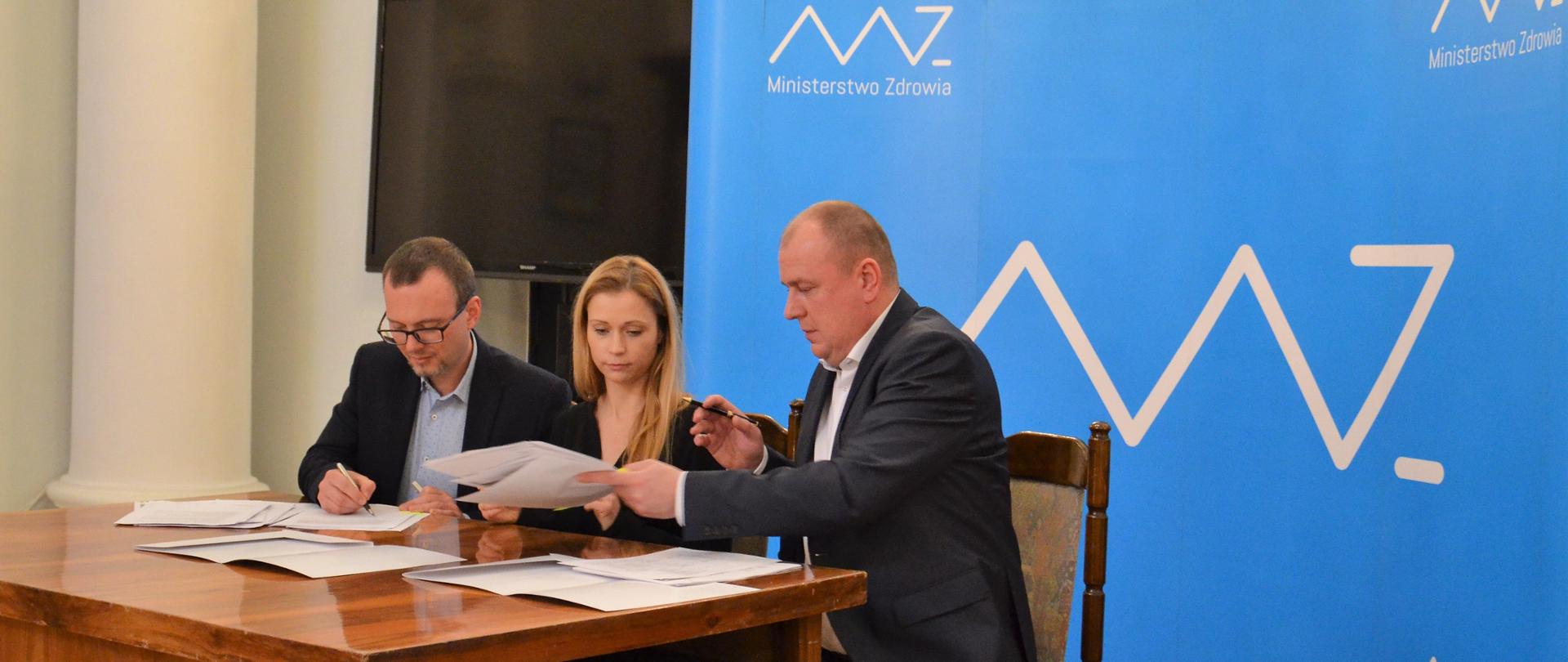 Podpisanie umów w siedzibie Ministerstwa Zdrowia. Przy stole siedzą trzy osoby, przedstawiciele beneficjentów oraz zastępca dyrektora Departamentu Funduszy Europejskich i e-Zdrowia Anna Goławska.