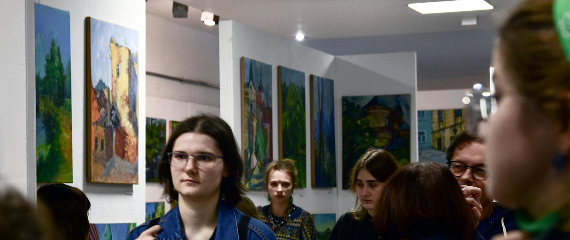 Uczniowie oglądają wystawę malarstwa w holu szkoły. Na ścianach są zawieszone obrazy.