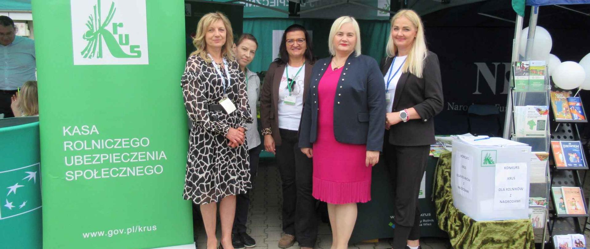 Pięć uśmiechniętych kobiet stoi przed zielonym namiotem z napisem „PRACUJ BEZPIECZNIE” i banerem KRUS.