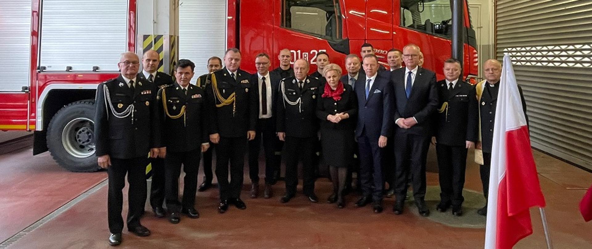 Fotografia wykonana w garażu jednostki ratowniczo-gaśniczej nr 1 komendy powiatowej Państwowej Straży Pożarnej w Mielcu. Grupowe zdjęcie zaproszonych gości i funkcjonariuszy. Funkcjonariusze ubrani w galowe mundury koloru granatowego Państwowej Straży Pożarnej. Zdjęcie wykonane na tle samochodu ratowniczo-gaśniczego przekazanego komendzie powiatowej Państwowej Straży Pożarnej w Mielcu.