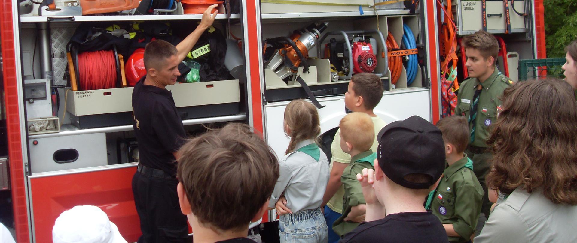 Na wprost w tle samochód strażacki z otwartymi skrytkami gdzie widać sprzęt będący na wyposażeniu strażackiego wozu, strażak pokazujący sprzęt, na 1 planie stojące tyłem dzieci biorące udział w spotkaniu. 