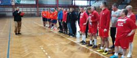 Zdjęcie dokumentuje oficjalne zkończenie wydarzenia sportowego w hali, uchwyconą z perspektywy bocznej, która podkreśla liczebność uczestników. Po lewej stronie kadru stoi mężczyzna w czarnej kurtce mundurowej i spodniach bojówkach. W dłoniach trzyma dyplom oraz mikrofon, co sugeruje, że odczytuje wyniki zawodów lub wygłasza przemówienie końcowe. Przed nim, wzdłuż jednej z linii boiska, ustawione są w szeregu dwie główne drużyny: Drużyna w czerwonych strojach (bliżej obiektywu): Grupa mężczyzn reprezentująca KP PSP Wołów. Niektórzy zawodnicy mają na sobie dresowe bluzy, inni koszulki z krótkim rękawem. Uwagę zwracają detale, takie jak białe getry i różnorodne obuwie sportowe (np. jaskrawożółte i niebieskie buty halowe). Jeden z zawodników w środku szeregu trzyma dużą butelkę wody. Drużyna w pomarańczowych strojach (dalej w szeregu): Zawodnicy w jaskrawych koszulkach stoją w rzędzie za grupą w czerwieni. Pomiędzy obiema drużynami stoją osoby w ubraniach cywilnych lub dresach (m.in. w niebieskiej bluzie).