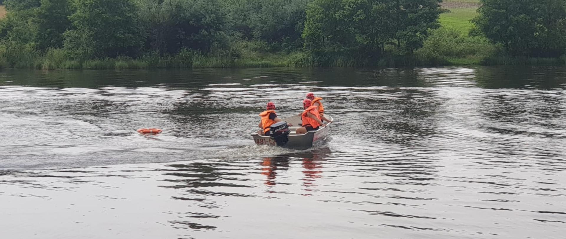 Na zdjęciu widzimy strażaków ubranych w kamizelki ratunkowe płynących łodzią ratowniczą do poszkodowanego w celu wyciągnięci ago. Za poszkodowanego służby koło ratunkowe. W tle widać drzewa.
