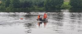 Na zdjęciu widzimy strażaków ubranych w kamizelki ratunkowe płynących łodzią ratowniczą do poszkodowanego w celu wyciągnięci ago. Za poszkodowanego służby koło ratunkowe. W tle widać drzewa.