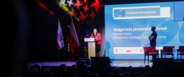 Na scenie przemawia Sekretarz Stanu Małgorzata Jarosińska-Jedynak. W tle widać ekran konferencji. Po prawej stronie stoi mężczyzna.