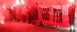 Ludzie odpalają race ora trzymają duży banner z napisem Gostyń.