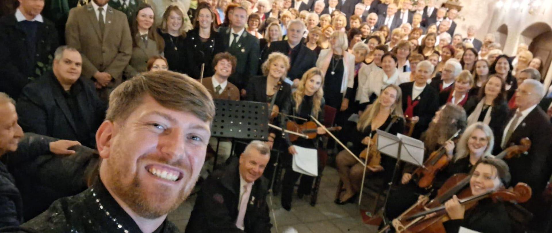 Nauczyciel chóru i orkiestry Dominik Jarocki pozuje do zdjęcia grupowego z wszystkimi wykonawcami koncertu w ramach XXIV Międzynarodowego Festiwalu im. Ignazego Reimanna w Krosnowicach , który odbył się 27 września 2025 