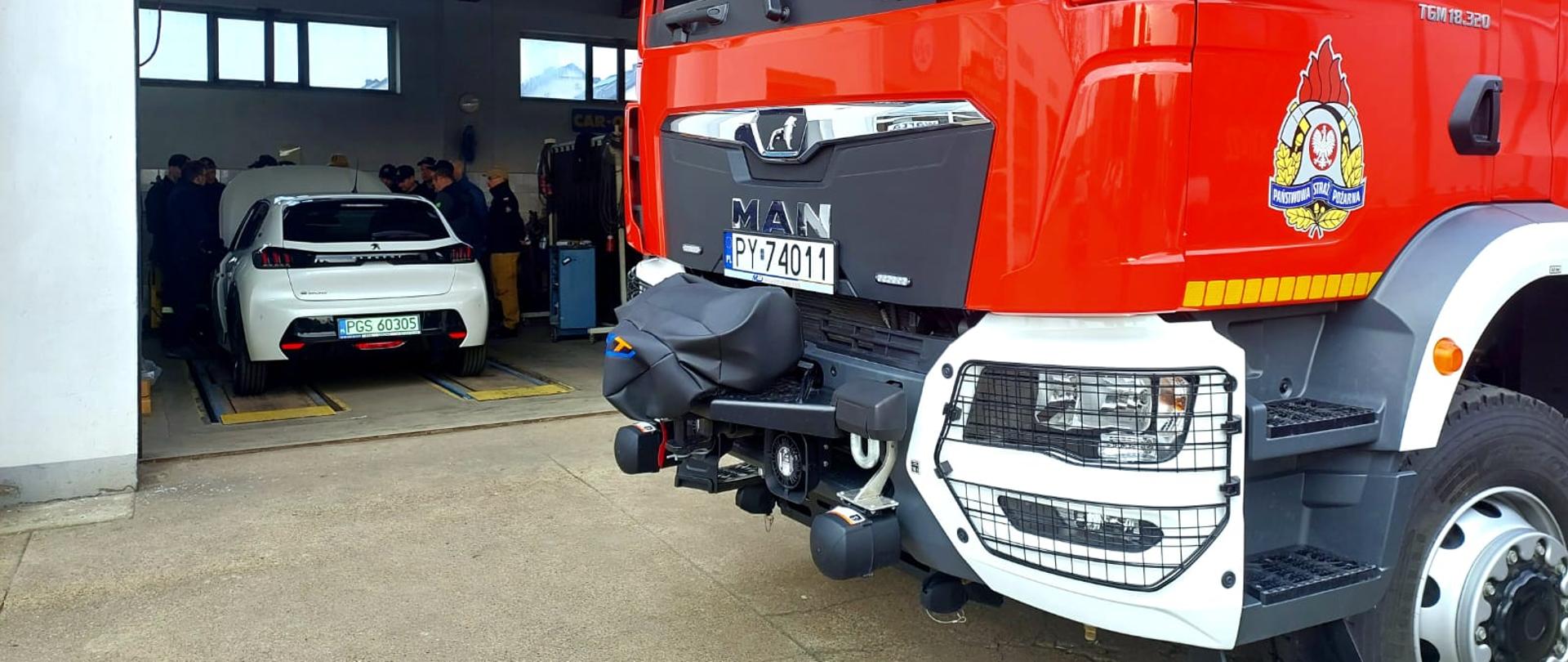 Strażaków którzy stoją przy samochodzie elektrycznym. Samochód znajduje się w garażu. Przed garażem stoi wóz strażacki.