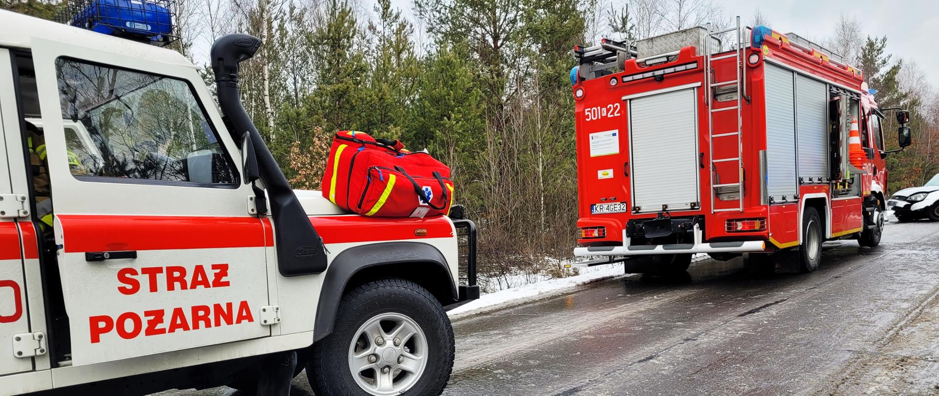 Wypadek samochodu osobowego, na zdjęciu samochód operacyjny biały oraz samochód bojowy straży pożarnej czerwony w tle uszkodzony samochód osobowy