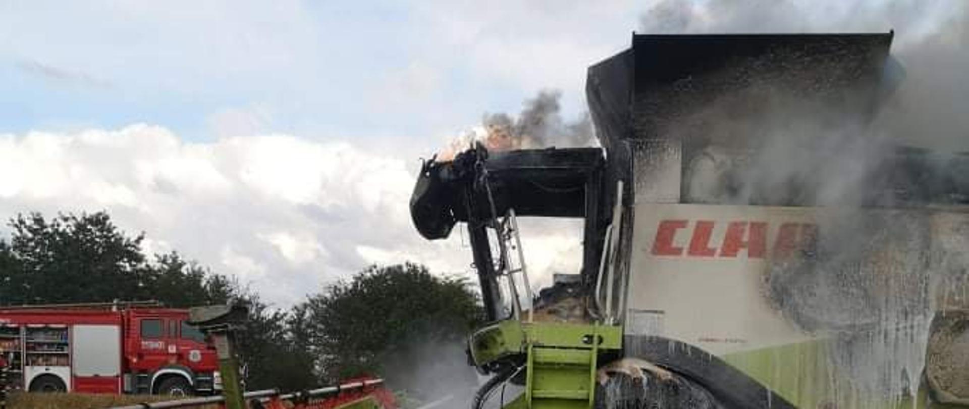Na zdjęciu widać pożar kombajnu zbożowego, który zapalił się podczas pracy na polu. W tle widać samochód straży pożarnej.