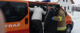 Zdjęcie zrobione na tle samochodu pożarniczego, typu bus. Przy samochodzie dwie osoby wchodzące do pojazdu poprzez boczne, otwarte, przesuwne drzwi oraz strażak w umundurowaniu specjalnym.