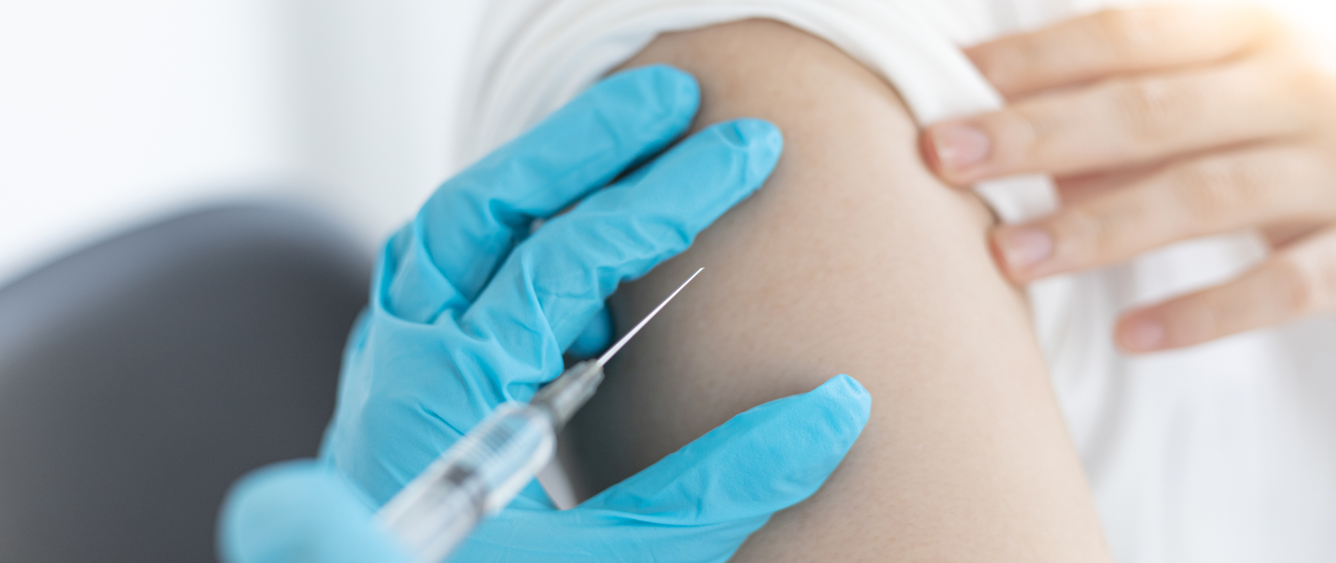 Na zdjęciu widzimy dłoń w niebieskiej jednorazowej rękawiczce trzymającą strzykawkę z igłą. Igła wbijana jest w ramię drugiej osoby.