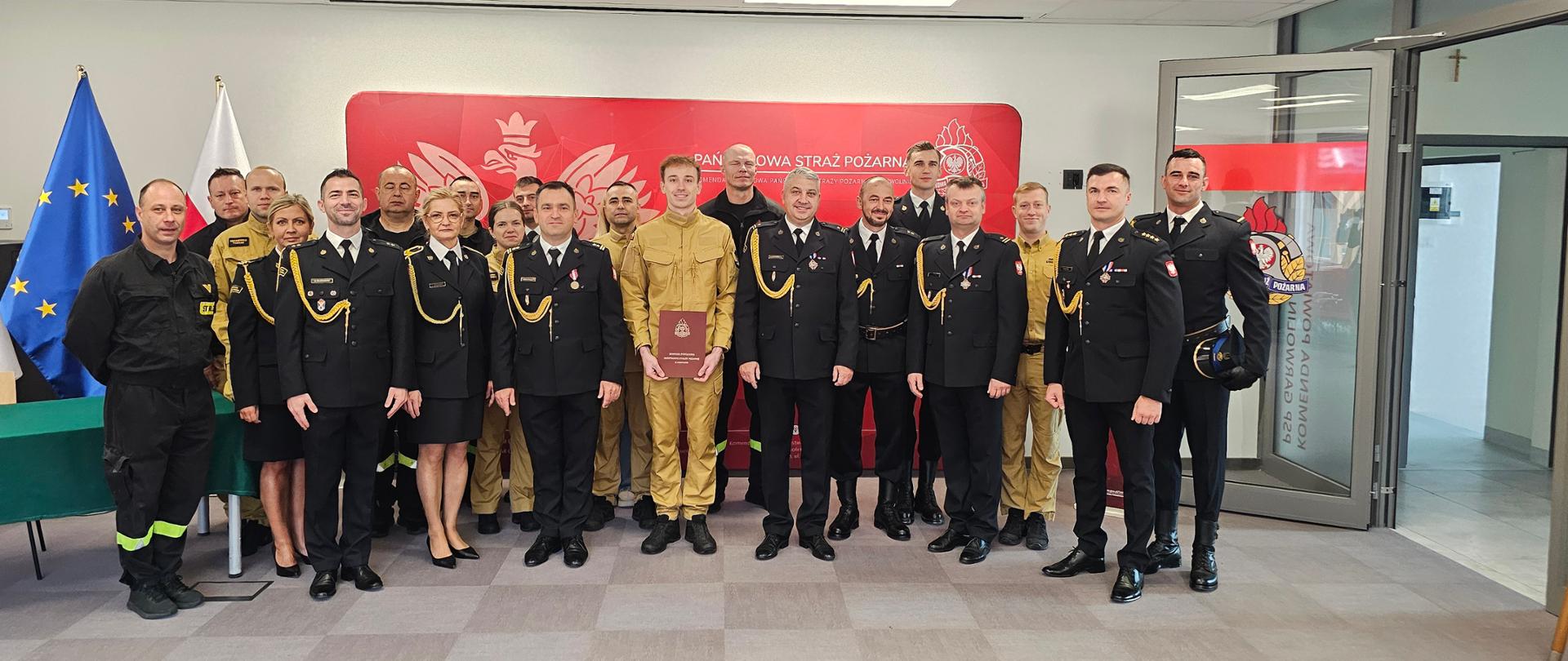 Na zdjęciu grupowym w sali komendy pozują funkcjonariusze Państwowej Straży Pożarnej oraz goście zgromadzeni wokół wyróżnionego strażaka w piaskowym mundurze. Tło tworzy czerwona ścianka reprezentacyjna z godłem Polski oraz stojące obok flagi państwowe i Unii Europejskiej.