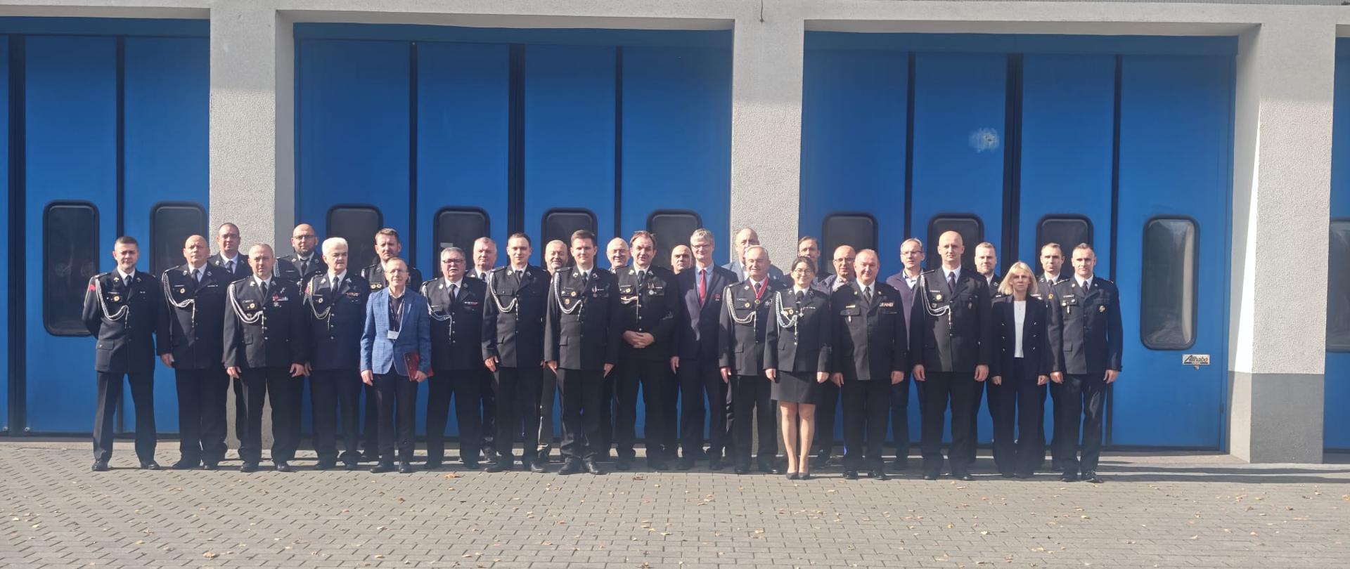 Strażacy w ubraniach wyjściowych stoją na zbiórce do zdjęcia przed niebieskimi bramami komendy.