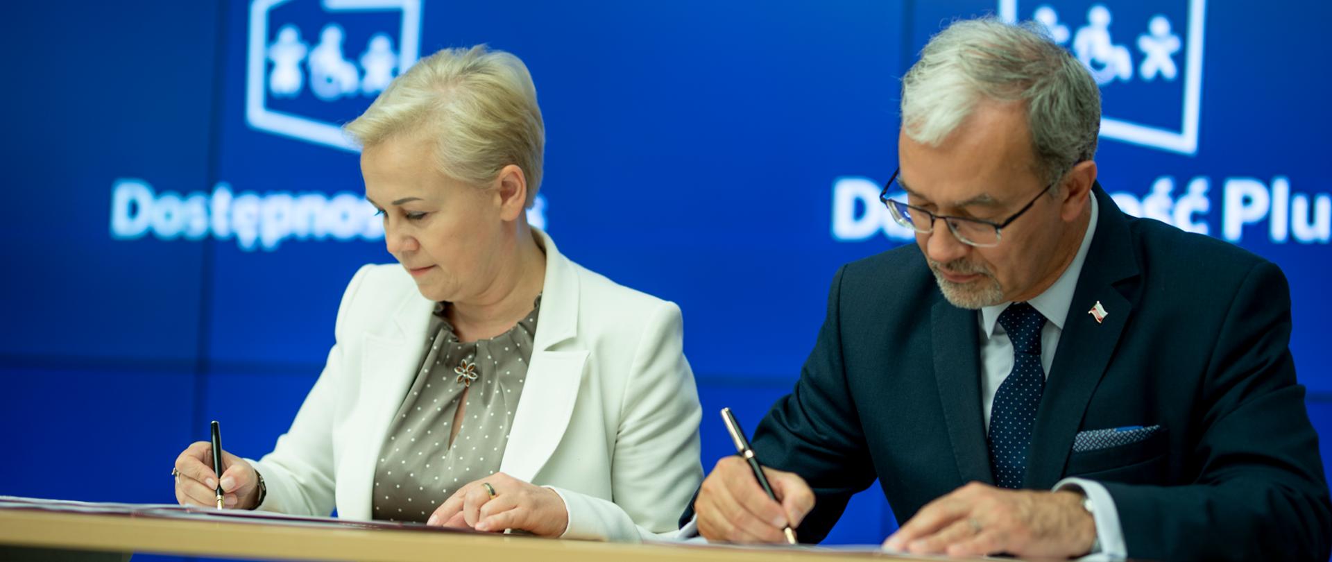 Minister inwestycji i rozwoju Jerzy Kwieciński i prezes zarządu Banku Gospodarstwa Krajowego Beata Daszyńska-Muzyczka podpisują umowę. Za nimi na ekranie logotyp programu Dostępność Plus.