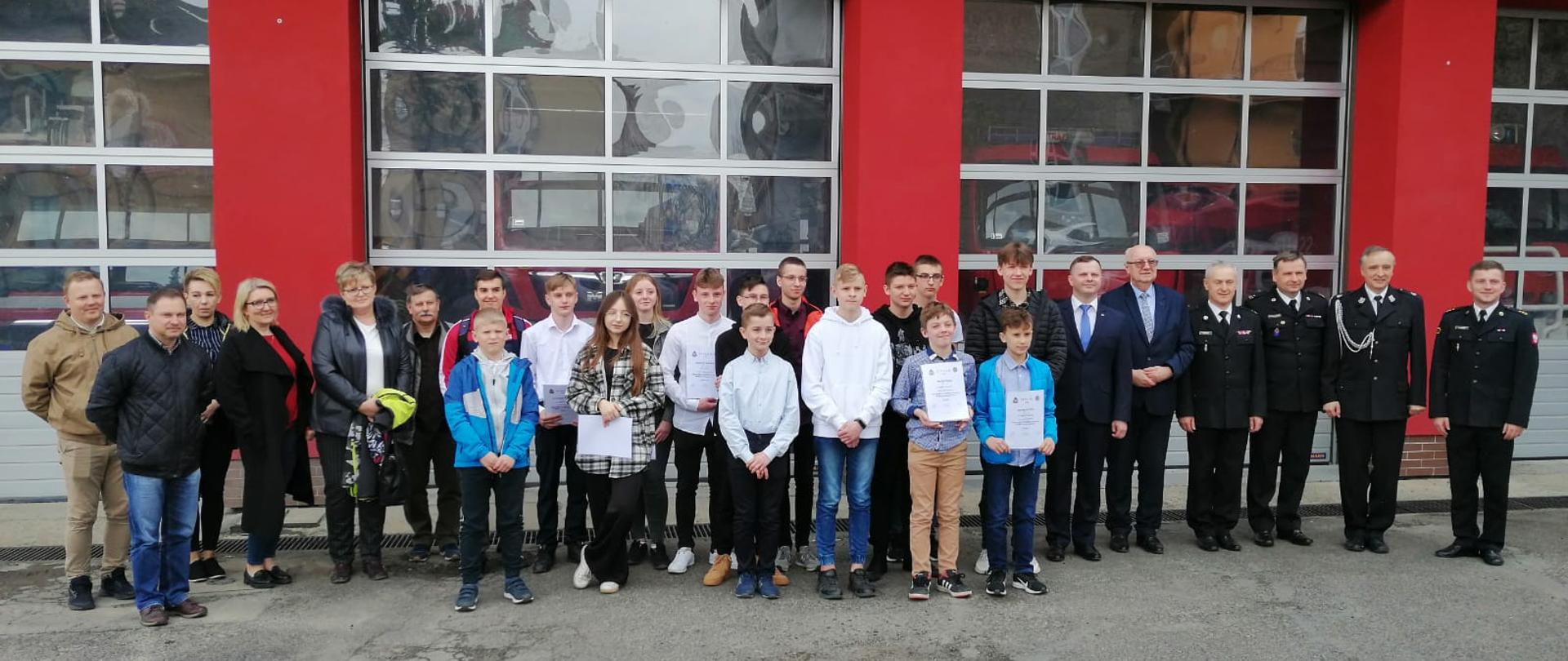 Kolorowa fotografia wykonana na zewnątrz przed czerwonym budynkiem straży pożarnej z bramami wyjazdowymi. Przed budynkiem stoi grupa osób cywilnych dorosłych oraz młodzieży z dyplomami. Obok stoją też umundurowani strażacy. Na budynku widoczna część srebrnego napisu STRAŻ POŻARNA. Zdjęcie wspólne uczestników eliminacji powiatowych 2022 Ogólnopolskiego Turnieju Wiedzy Pożarniczej "Młodzież Zapobiega Pożarom".