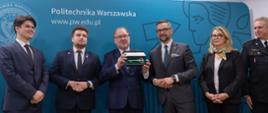 Pod niebieską ścianką z napisem Politechnika Warszawska stoi w rzędzie sześć osób, pośrodku mężczyzna w garniturze pokazuje otwarte pudełko z dużym ozdobnym kluczem, obok niego stoi minister Kulasek.