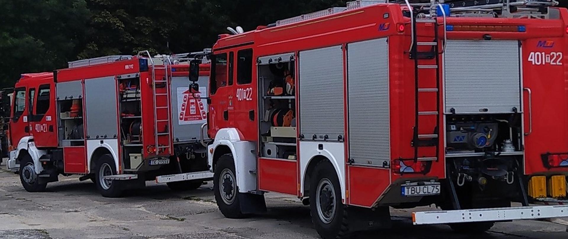 2 samochody pożarnicze koloru czerwonego stojące jeden za drugim. W tle drzewa.