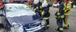 Strażacy wykonują cięcie nożycami hydraulicznymi na wraku samochodu osobowego. Samochód koloru niebieskiego. W tle ustawiony bokiem samochód pożarniczy. Inni strażacy stoją obok ćwiczących. 