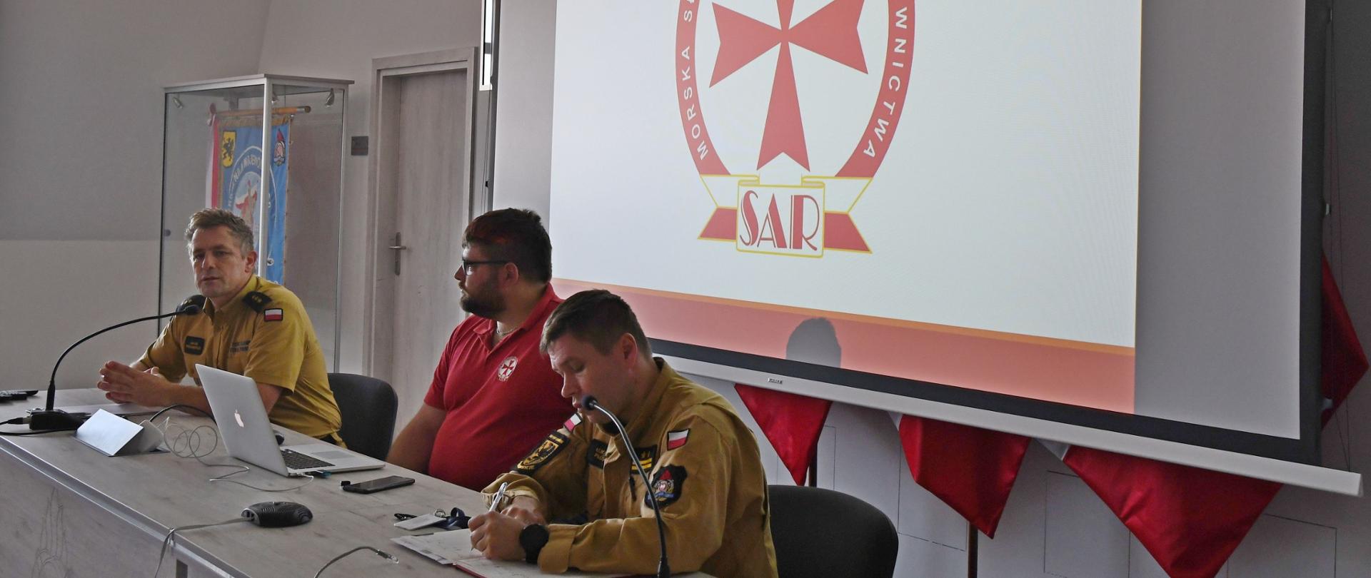 Dwóch strażaków Państwowej Straży Pożarnej oraz przedstawiciel Morskiej Służby Poszukiwania i Ratownictwa siedzą za biurkiem na którym rozłożony jest laptop stoją mikrofony za nimi na ekranie wyświetlana jest prezentacja Morska Służba Poszukiwania i Ratownictwa logo SAR.