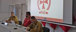 Dwóch strażaków Państwowej Straży Pożarnej oraz przedstawiciel Morskiej Służby Poszukiwania i Ratownictwa siedzą za biurkiem na którym rozłożony jest laptop stoją mikrofony za nimi na ekranie wyświetlana jest prezentacja Morska Służba Poszukiwania i Ratownictwa logo SAR.
