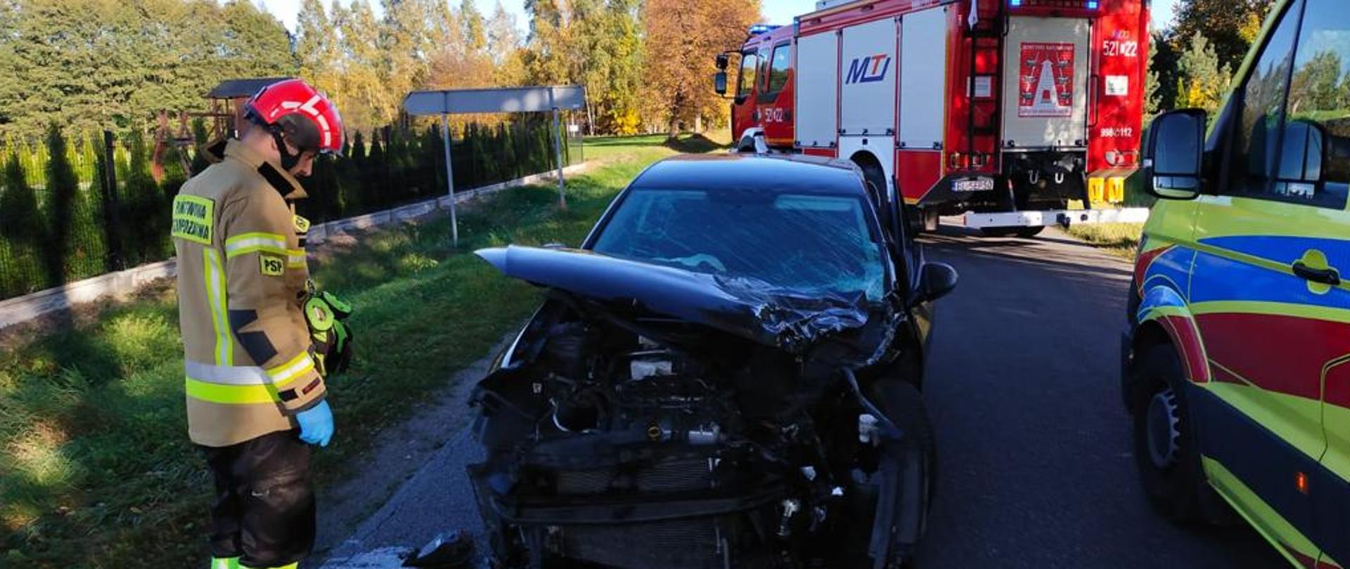 Na drodze asfaltowej stoi czarny samochód osobowy ze skasowanym przodem w wyniku zderzenia z innym samochodem. Po lewej stronie samochodu stoi strażak, a po prawej samochód karetki pogotowia. Za samochodem stoi samochód strażacki.