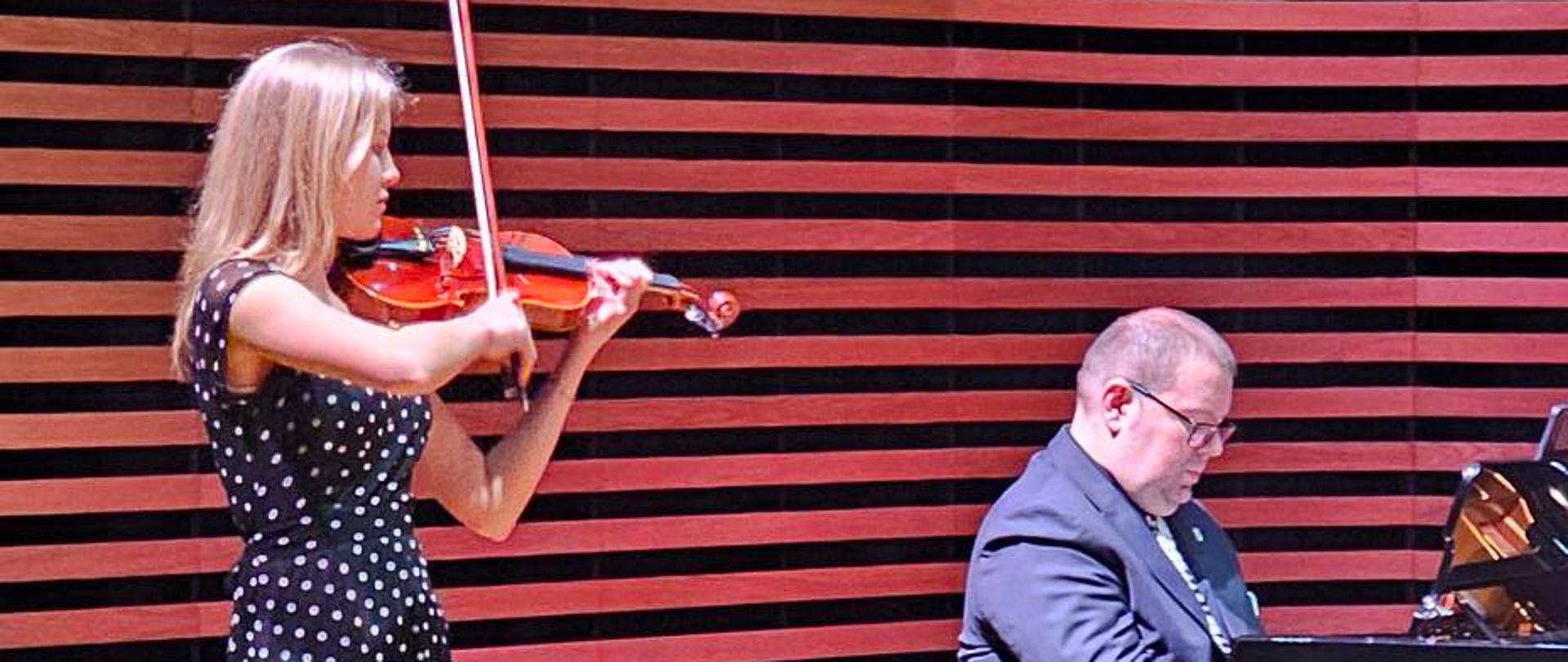 Dziewczyna gra na skrzypcach, a mężczyzna akompaniuje jej na fortepianie.