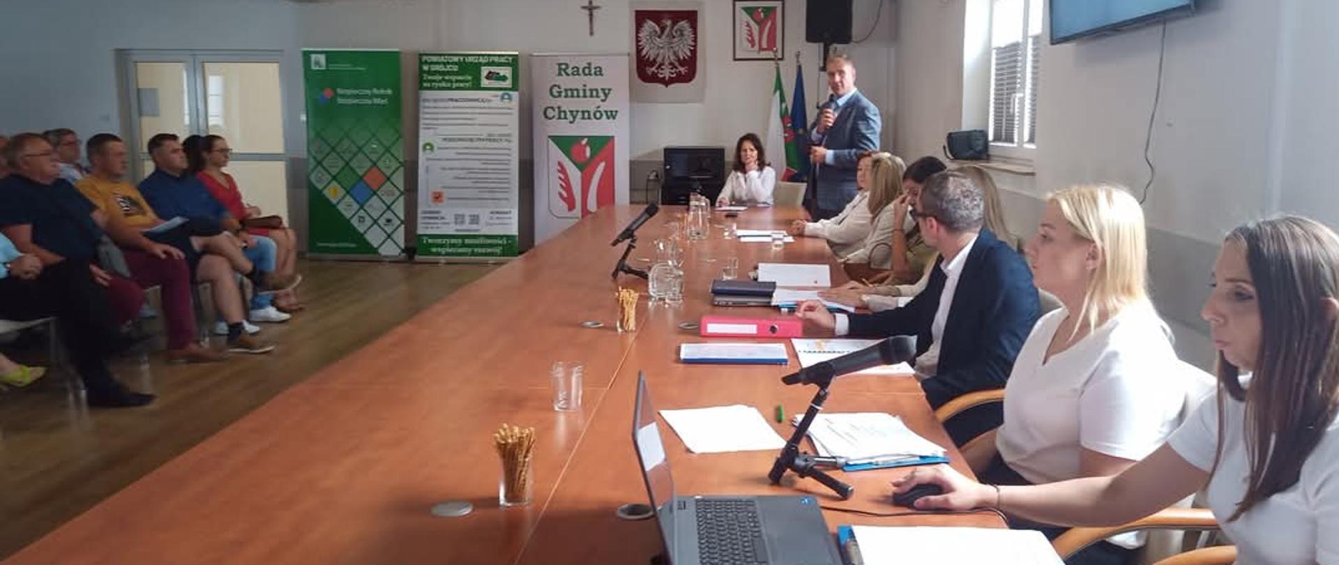 Mężczyzna z mikrofonem przemawia, obok za stołem siedzi grupa osób z notatkami. Przed nimi na krzesłach siedzą przysłuchujący się rolnicy.