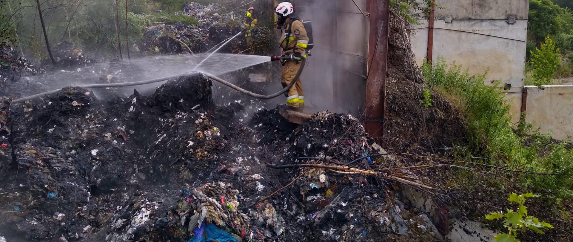 Hałda odpadów, częściowo spalona. Na hałdzie stoi strażak w ubraniu specjalnym w kolorze piaskowym, wyposażony w apart powietrzny, maskę oraz biały hełm. Strażak podaje na dymiącą się stertę odpadów prąd wody
