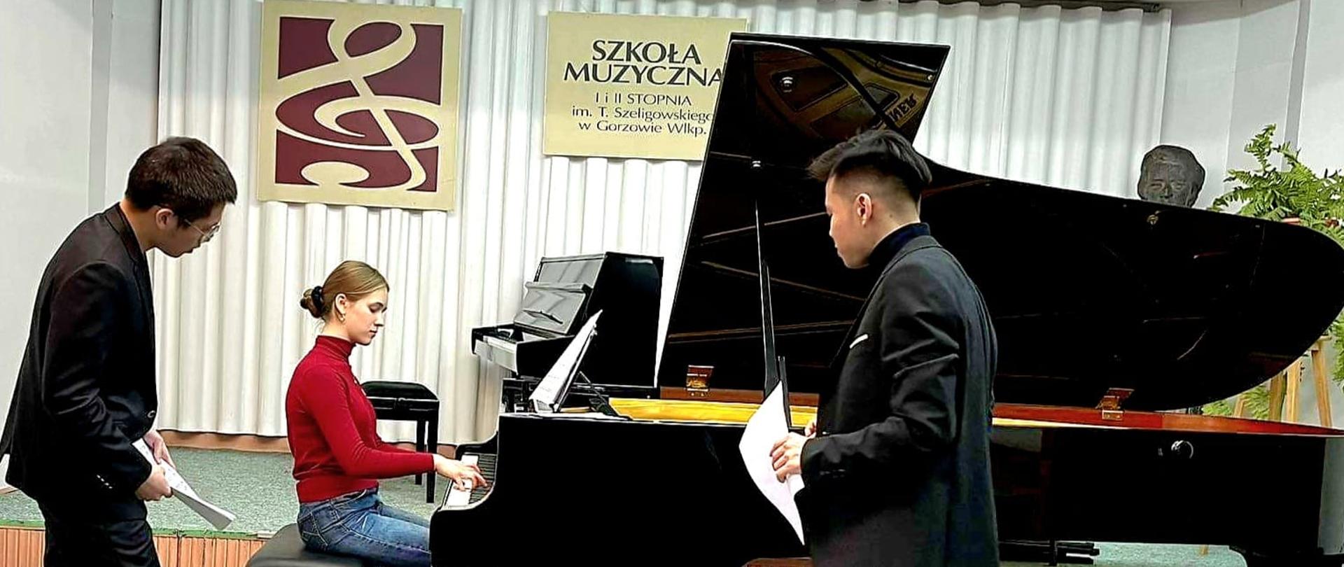 Zdjęcie: Przy czarnym fortepianie z otwartym skrzydłem siedzi uczennica w czerwonym swetrze i dżinsach, trzymając ręce na klawiaturze. W tle widać pianino. Na ścianie wisi grafika z kluczem wiolinowym i druga z nazwą szkoły. Po lewej i prawej stronie, przed fortepianem, stoją wykładowcy, patrzący się na uczennicę. Trzymają w rękach nuty.