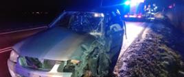 Zdjęcie przedstawia samochód osobowy koloru srebrnego. Samochód ma rozbity przód po zdarzeniu drogowym. W tle samochód strażacki z włączonymi światłami błyskowymi koloru niebieskiego.