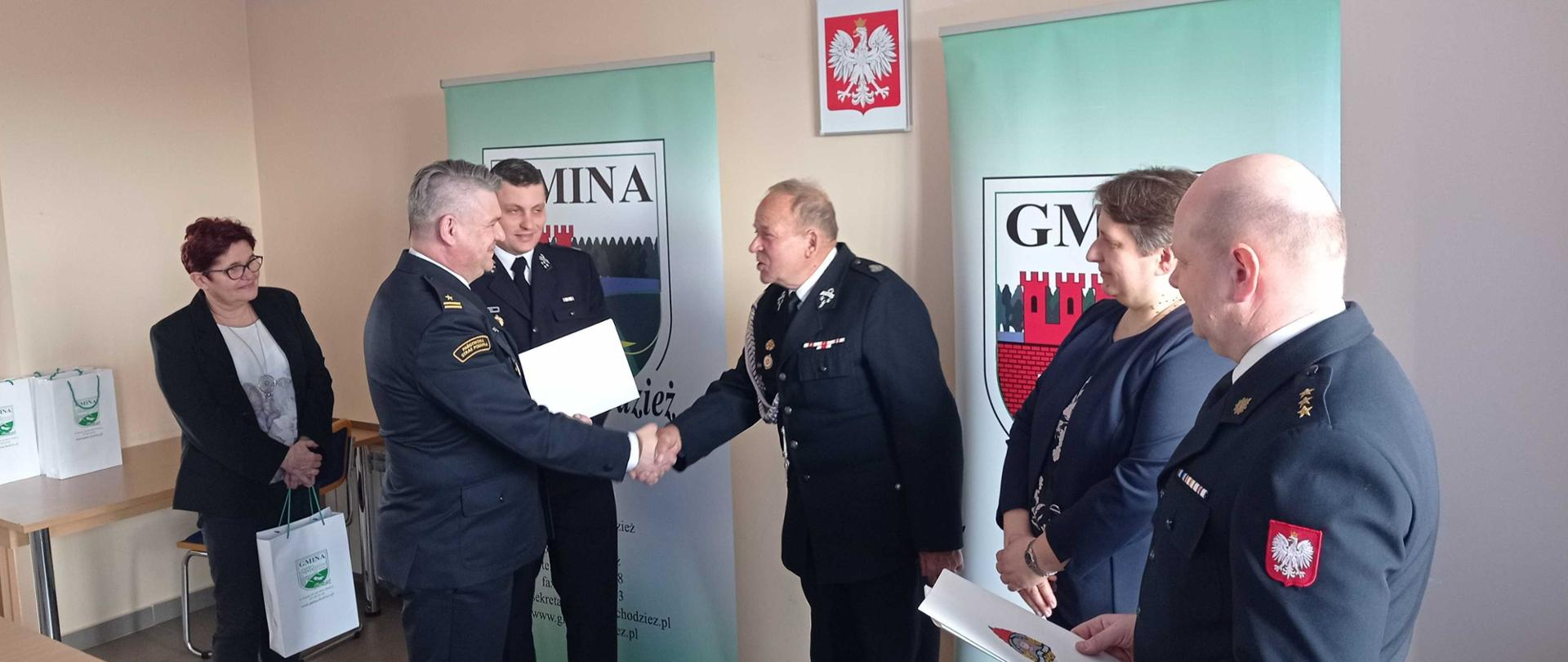 Zdjęcie przedstawia strażaków i samorządowców uczestniczących w uroczystym wręczeniu decyzjo świadczeń ratowniczych w pomieszczeniu w budynku Urzędu Gminy Chodzież.
W tle ROOL-UP Urzędu Gminy Chodzież.
