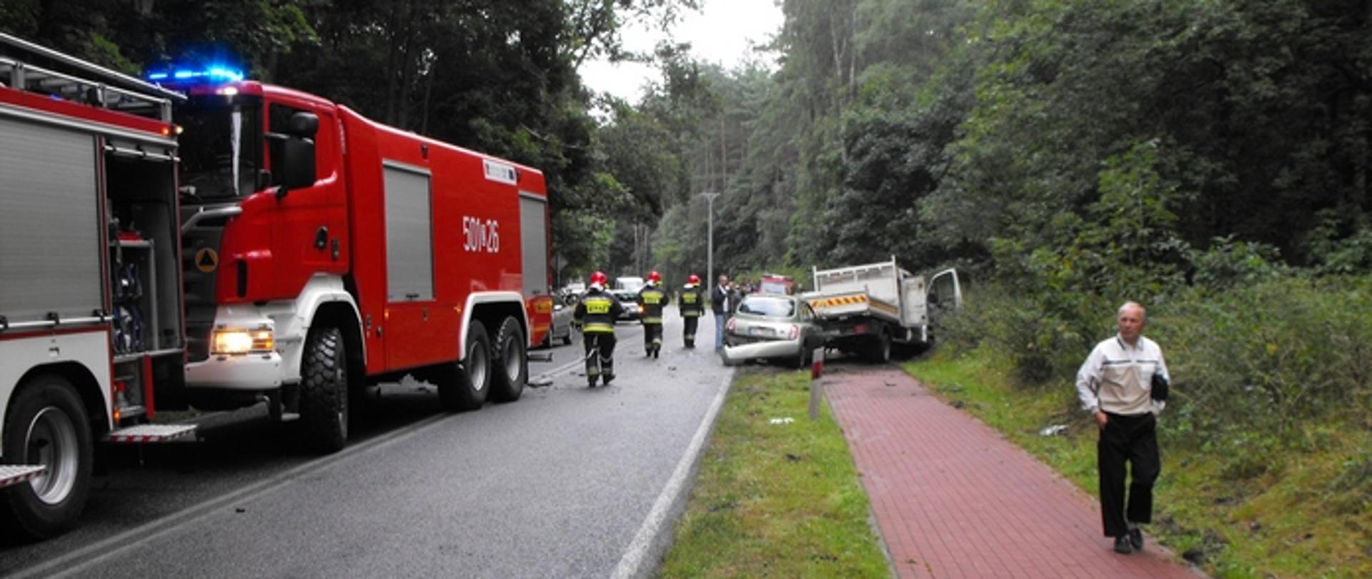 Zdjęcie przedstawia dwa rozbite samochody na ścieżce rowerowej oraz samochody pożarnicze na drodze