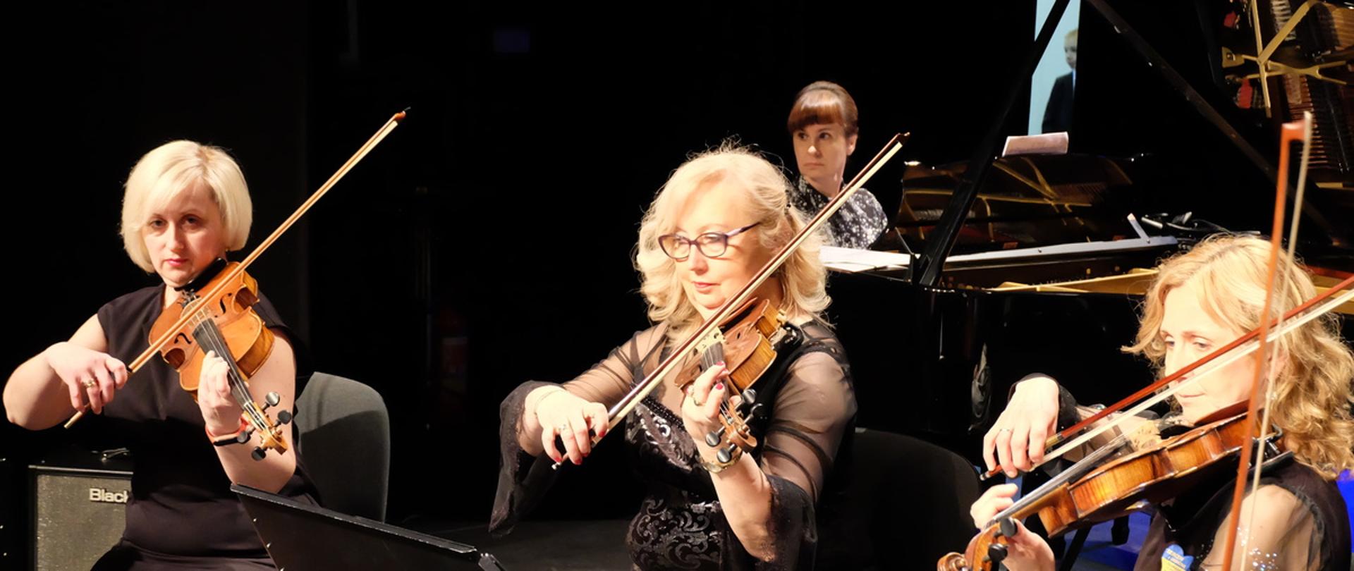 Trzy panie grają na skrzypcach. W tle przy fortepianie siedzi pianistka.