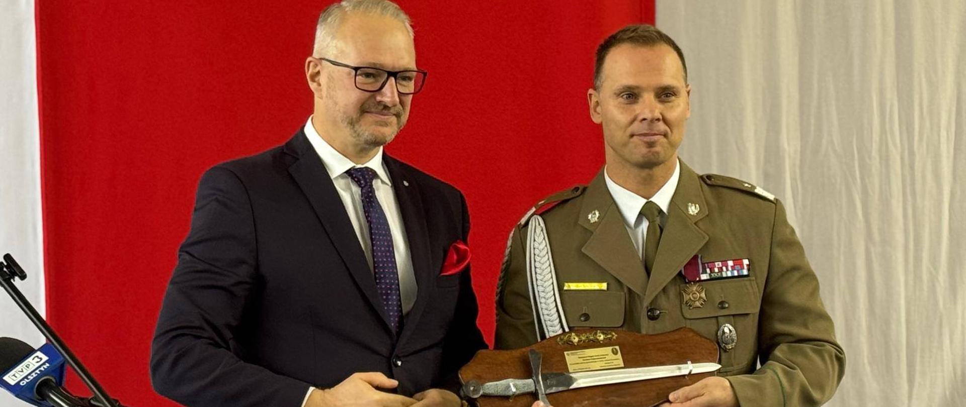 Dwóch mężczyzn stoi na czerwonym tle. Jeden z mężczyzn trzyma upominek w rękach 