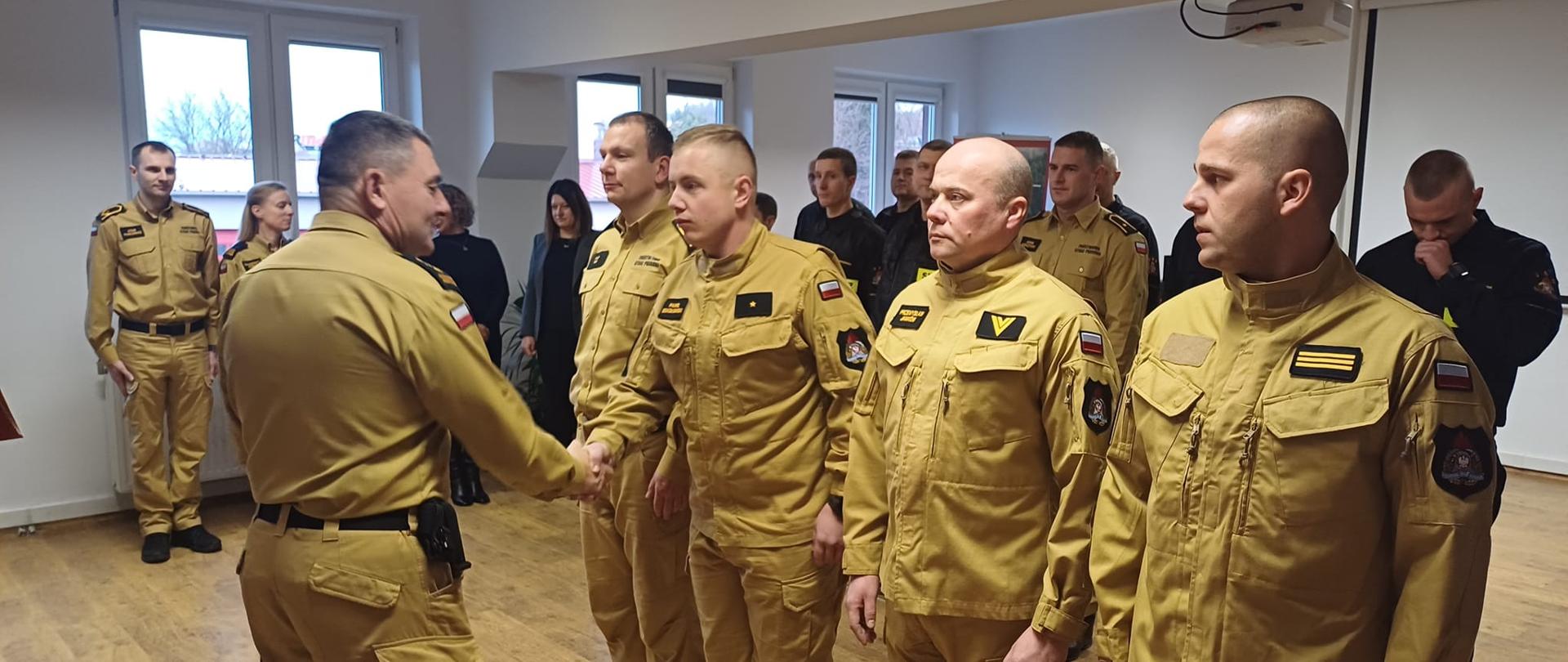 Na zdjęciu widoczni są funkcjonariusze Państwowej Straży Pożarnej ustawieni w szeregu podczas uroczystej zbiórki. Jeden z przełożonych wręcza gratulacje lub składa życzenia strażakowi poprzez uścisk dłoni, a pozostali funkcjonariusze oczekują na swoją kolej. W tle znajdują się inni uczestnicy uroczystości obserwujący przebieg wydarzenia.
