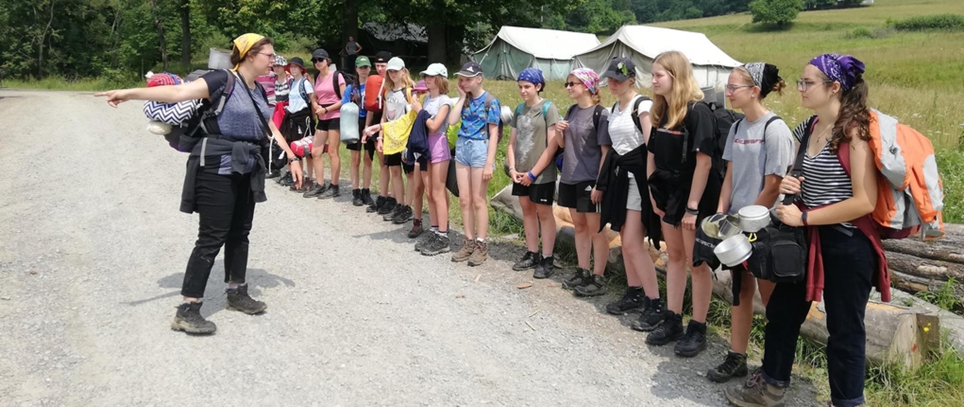 Na zdjęciu widocznych kilkunastu harcerzy stojących na zbiórce w szeregu podczas ewakuacji obozu harcerskiego. Harcerze stoją na szutrowej drodze za ich plecami łąka a w oddali las.