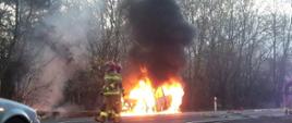 Na zdjęciu widać całkowity pożar samochodu osobowego, który stoi na poboczu drogi. Przed samochodem znajdują się dwaj strażacy natomiast w tle za palącym się samochodem jest las