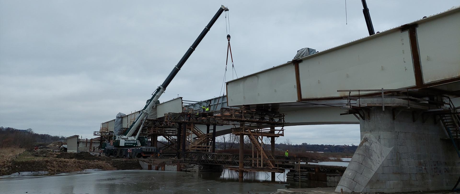 DK77 budowa mostu w Sandomierzu - montaż metalowych elementów konstrukcji przęsła z pomocą dźwigu. Pod mostem rzeka, w oddali brzeg, w wodzie betonowa podpora i podpory tymczasowe przypominające rusztowania. Na podporach szare stalowe podłużne elementy konstrukcji, miedzy nimi przerwa. W tym miejscu dźwig i pracownicy na rusztowaniu.